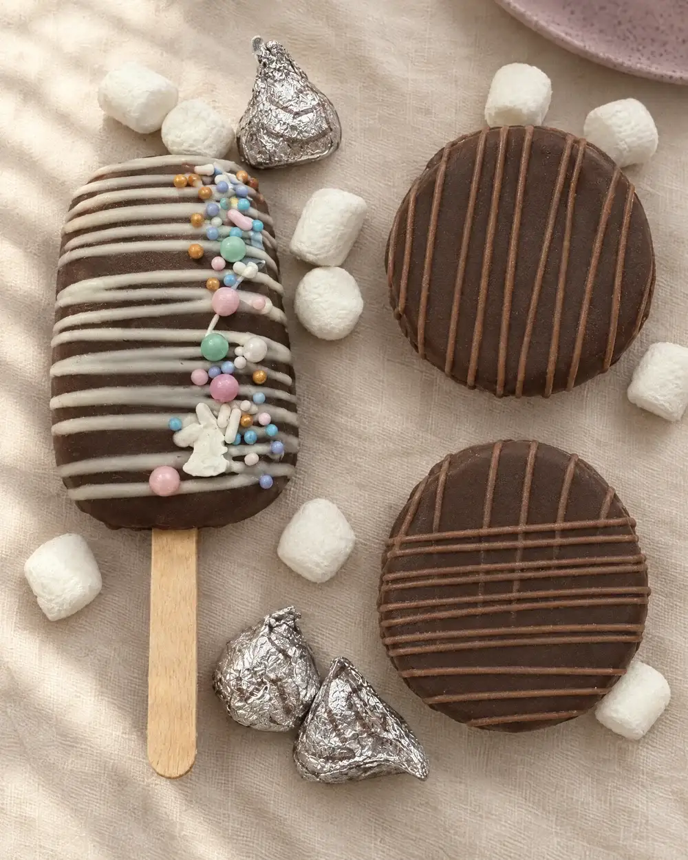 An assortment of cocoa bombs, a cakesicle, marshmallows and foil-wrapped chocolates on cream linen