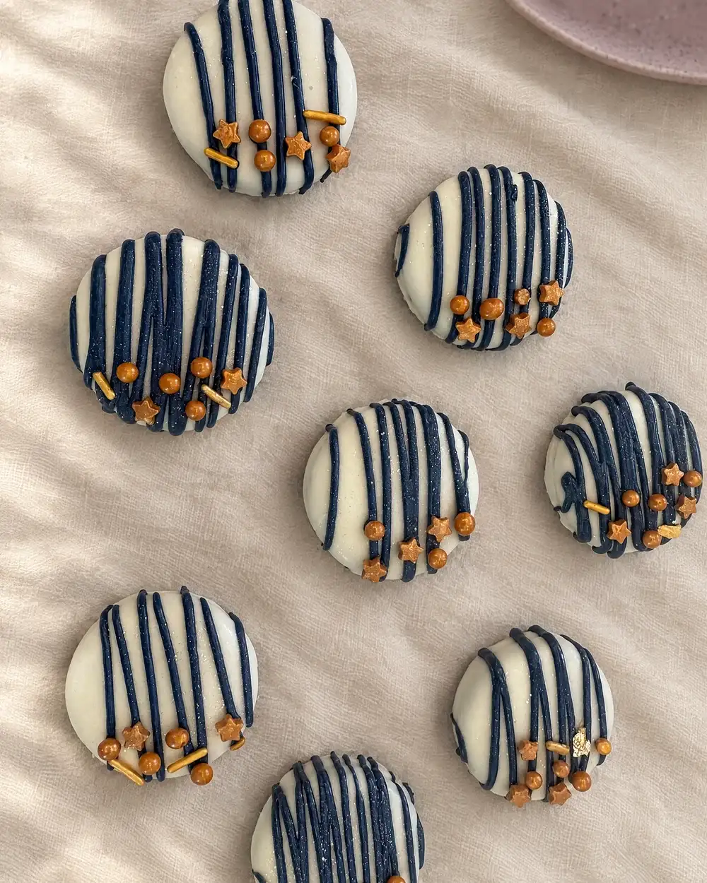 Eight chocolate-dipped Oreos in navy and white stripes with small gold stars, laid out on cream linen