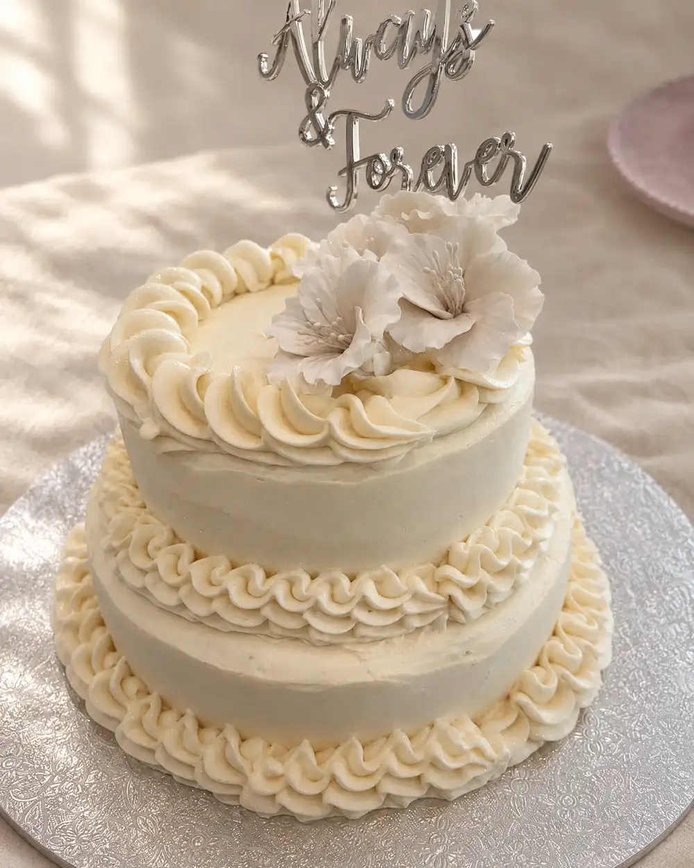 Two-tier ivory buttercream cake with sugar flowers and an Always and Forever topper, on cream linen